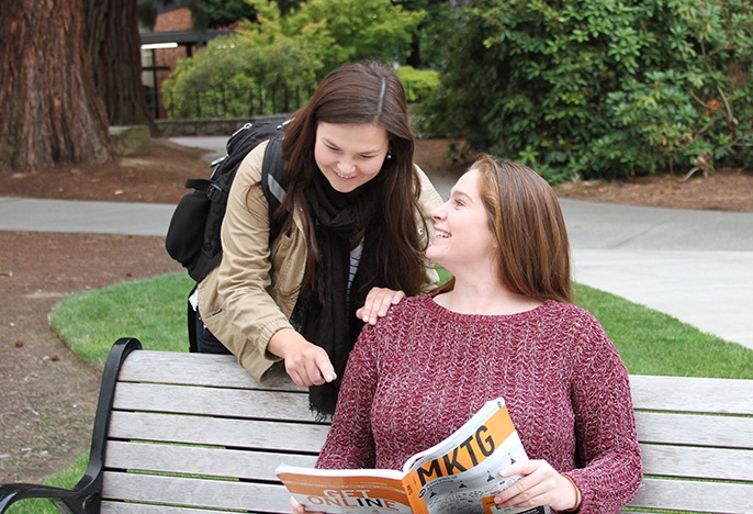 Two students discussing marketing outside on a bench.